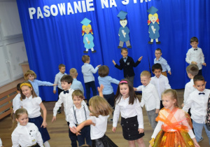 Na tle dekoracji dzieci ustawione w szachownicę tańczą żywiołowy taniec